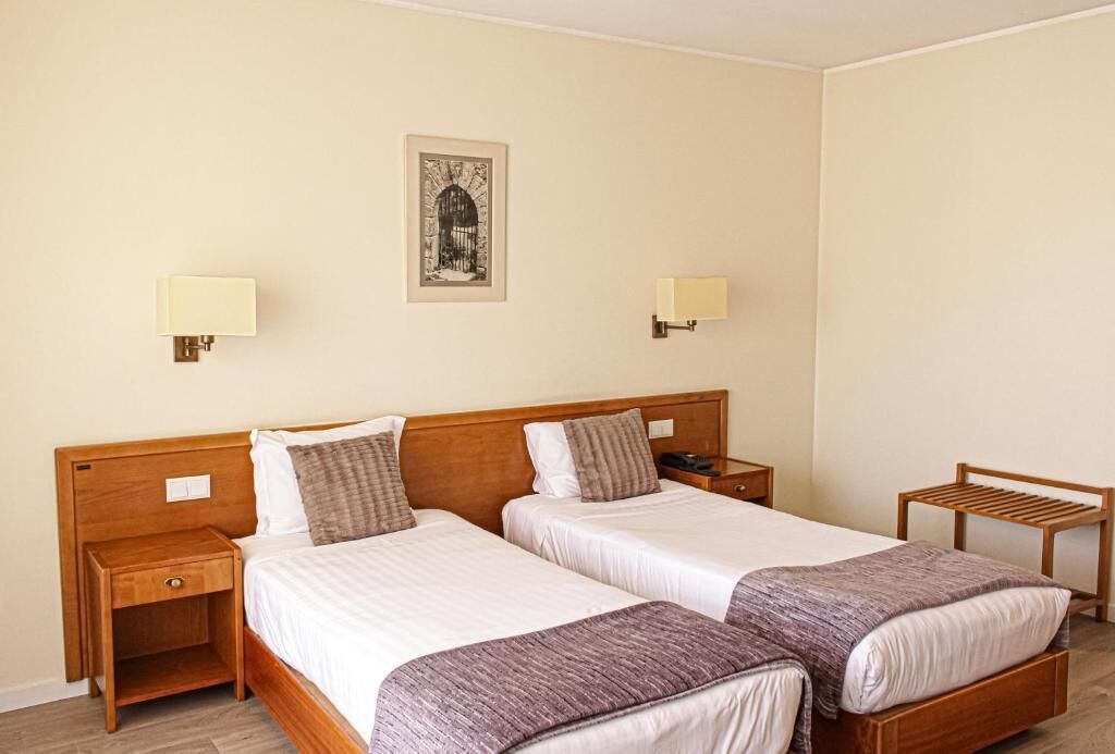 A hotel room with two single beds, wooden headboards, bedside tables with lamps, and a small wooden chair.