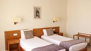 A hotel room with two single beds, wooden headboards, bedside tables with lamps, and a small wooden chair.