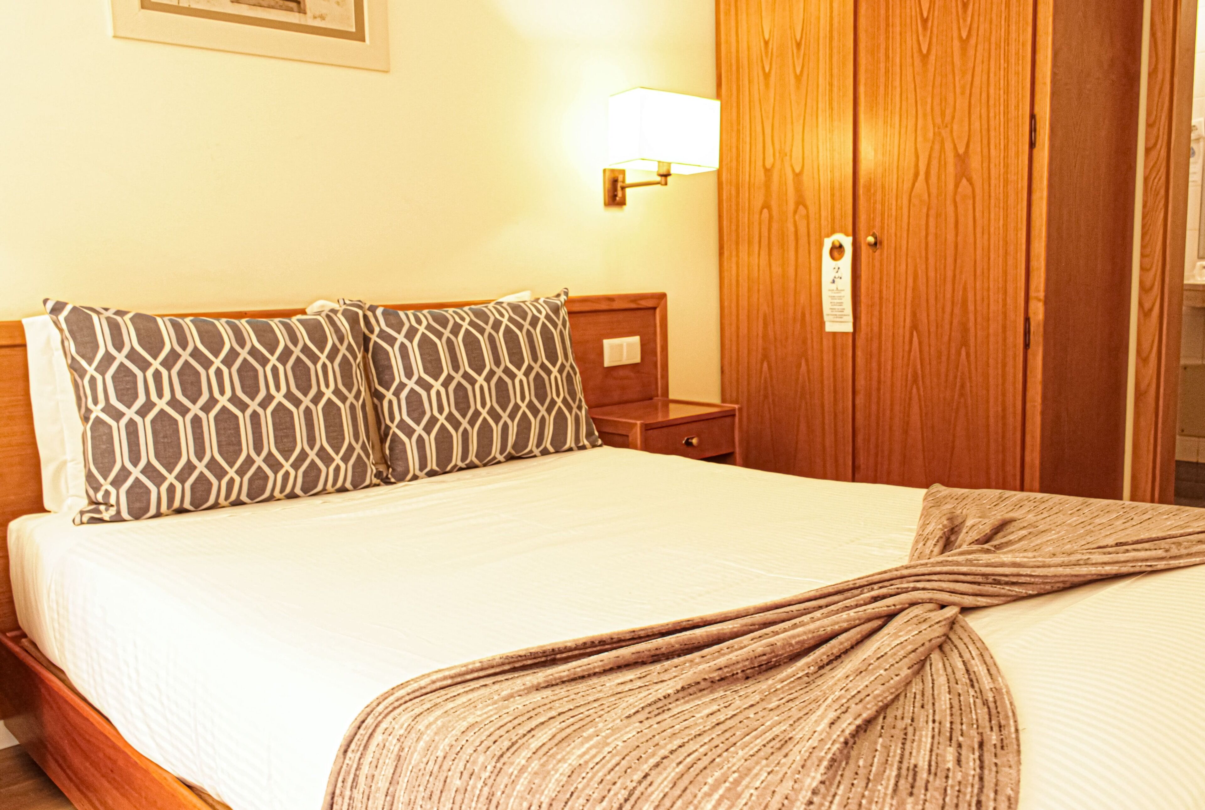 Una cama bien hecha con almohadas estampadas, una mesita de noche de madera y una lámpara de pared.