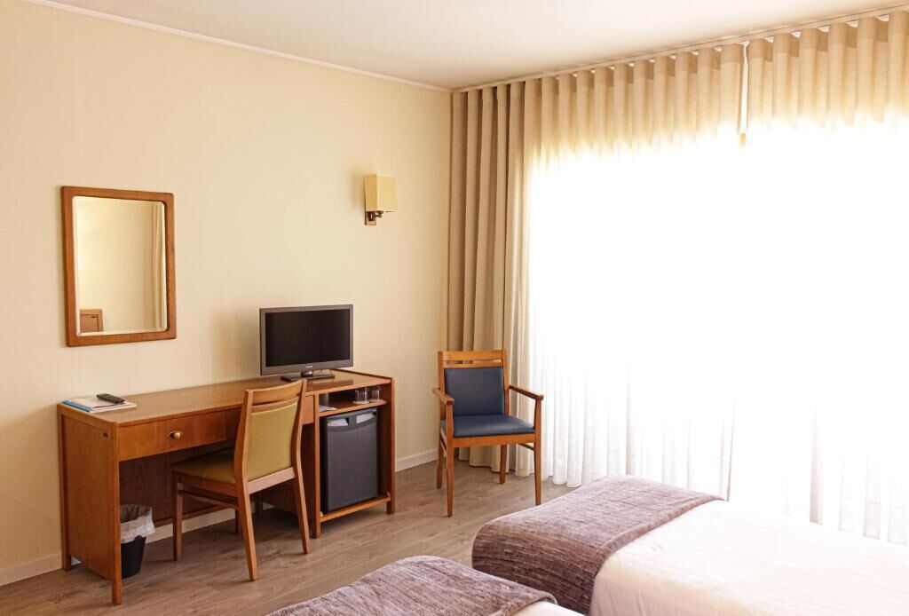 A hotel room with a bed, a desk with a computer, a chair, a television, and a window with curtains.