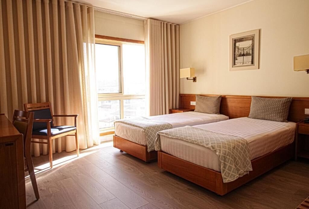 Une chambre d’hôtel avec deux lits, un bureau et une chaise. Il y a une fenêtre avec des rideaux, une lampe fixée au mur et un tableau encadré.