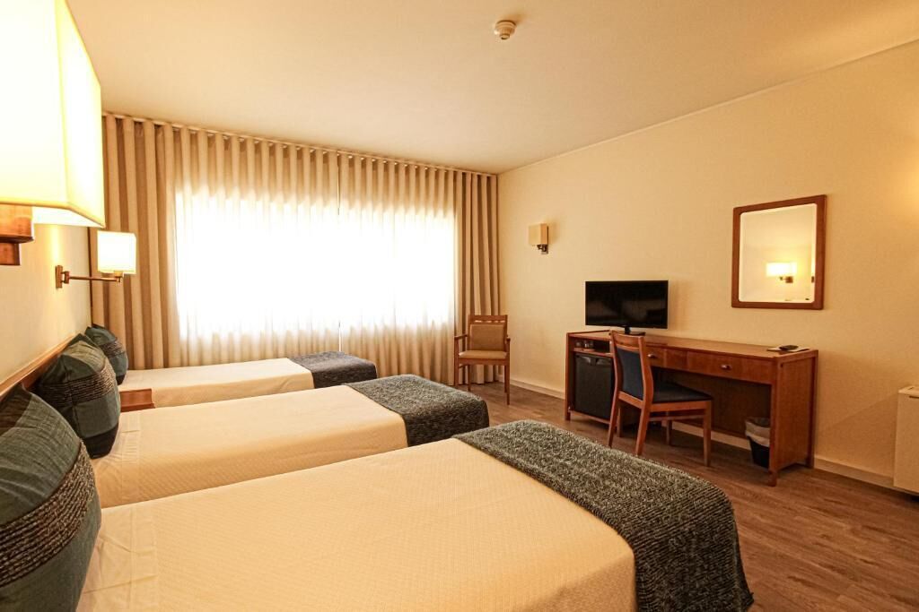 A hotel room with two single beds, a desk with a chair, a television, and a large window with curtains.