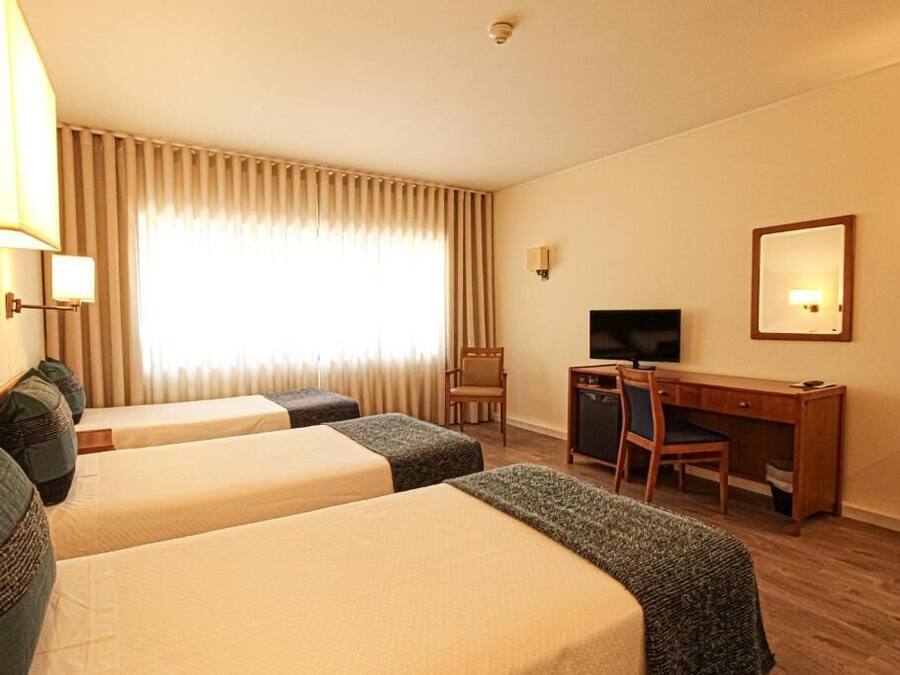 A hotel room with two single beds, a desk with a chair, a television, and a large window with curtains.