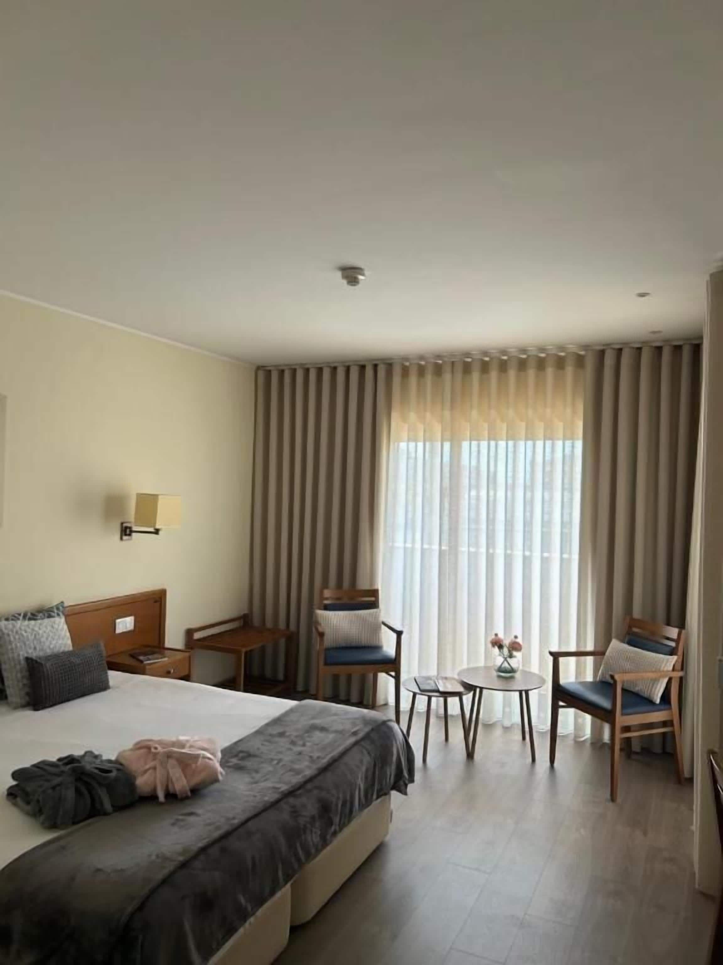 A hotel room with a bed, two chairs, a small table, and a window with curtains.