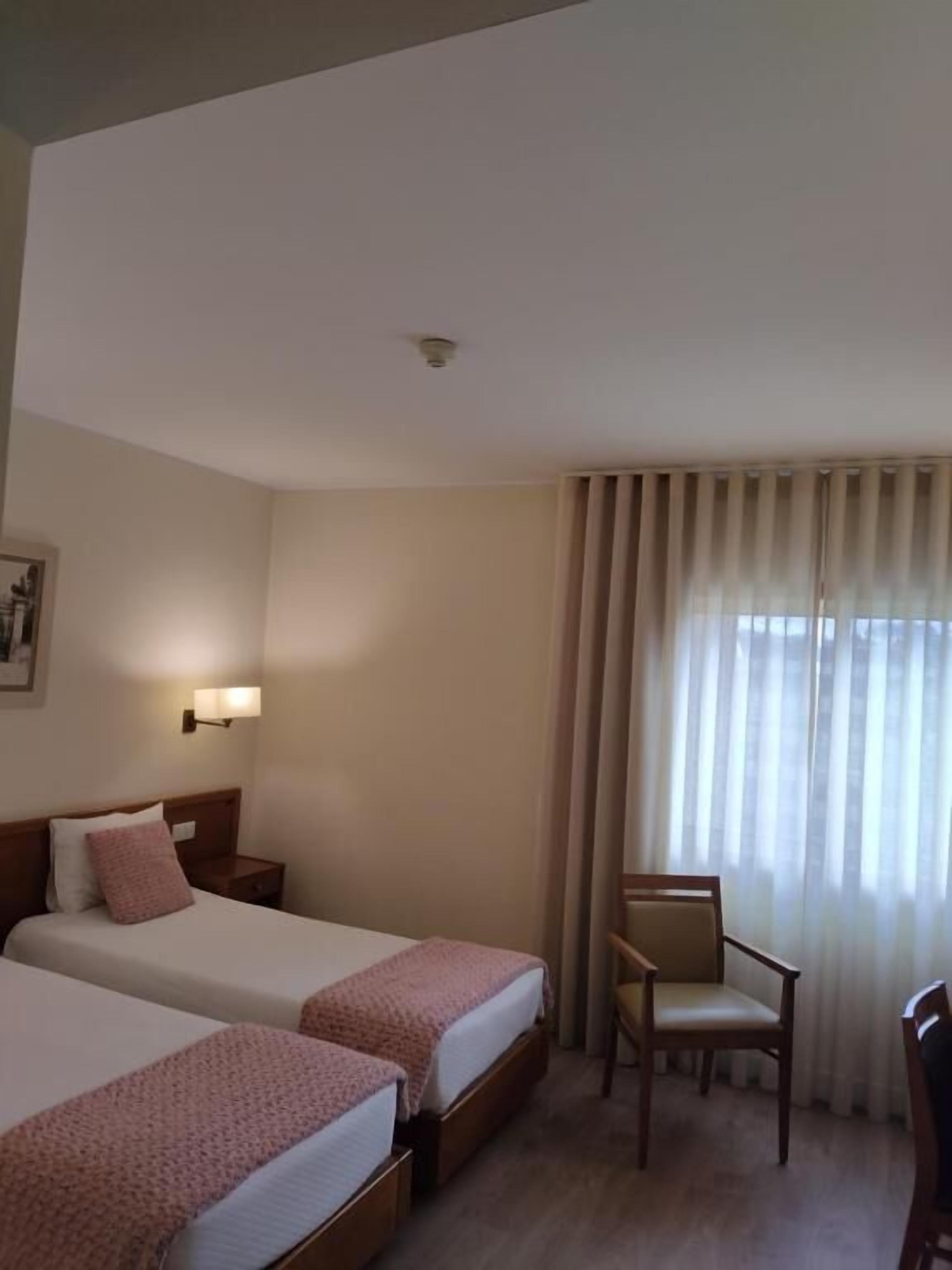 A hotel room with two beds, a chair, a nightstand, and a wall-mounted lamp.