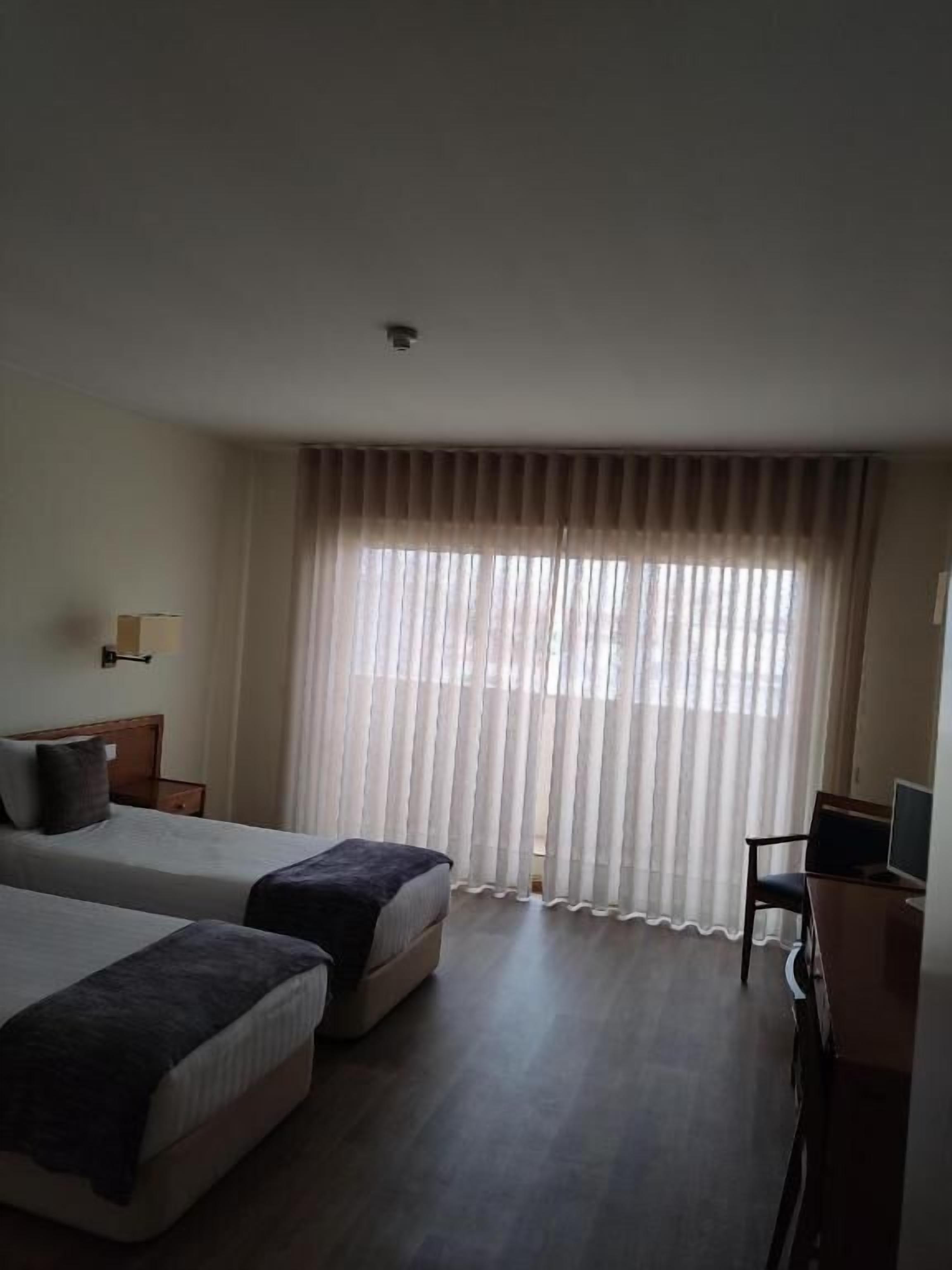 A hotel room with two beds, a desk with a chair, a television, and a large window with sheer curtains.