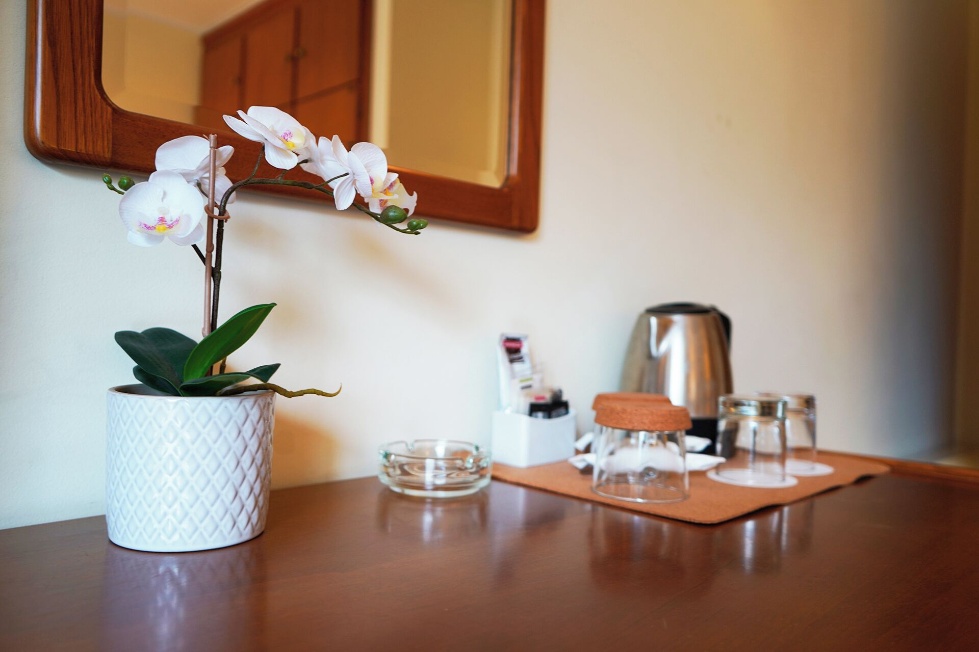 Un jarrón blanco texturizado con una orquídea sobre una mesa de madera, un pequeño cuenco de vidrio, un hervidor metálico y un vaso con tapón de corcho sobre un tapete de corcho.