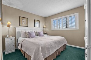 Une chambre avec un grand lit, deux tables de chevet, une fenêtre avec des stores et une moquette verte.
