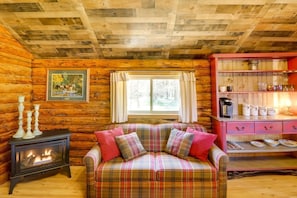 A cozy cabin interior with a plaid sofa, a wood-burning stove, and a shelf with kitchenware.