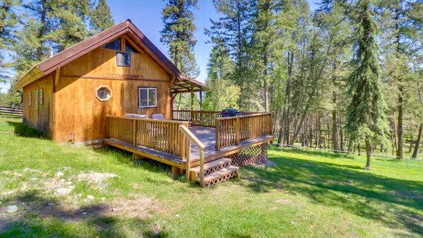 A wooden cabin with a deck, surrounded by tall trees. - Pet-Friendly Cottage w/ Hot Tub Lake Koocanusa (Rexford)