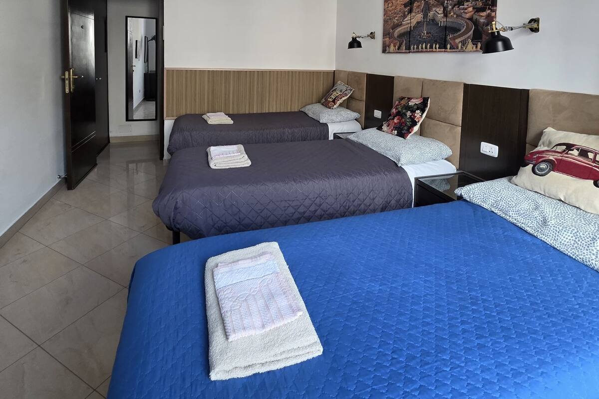 A hotel room with two beds, a wardrobe, a mirror, a wall-mounted lamp, and a cityscape painting.