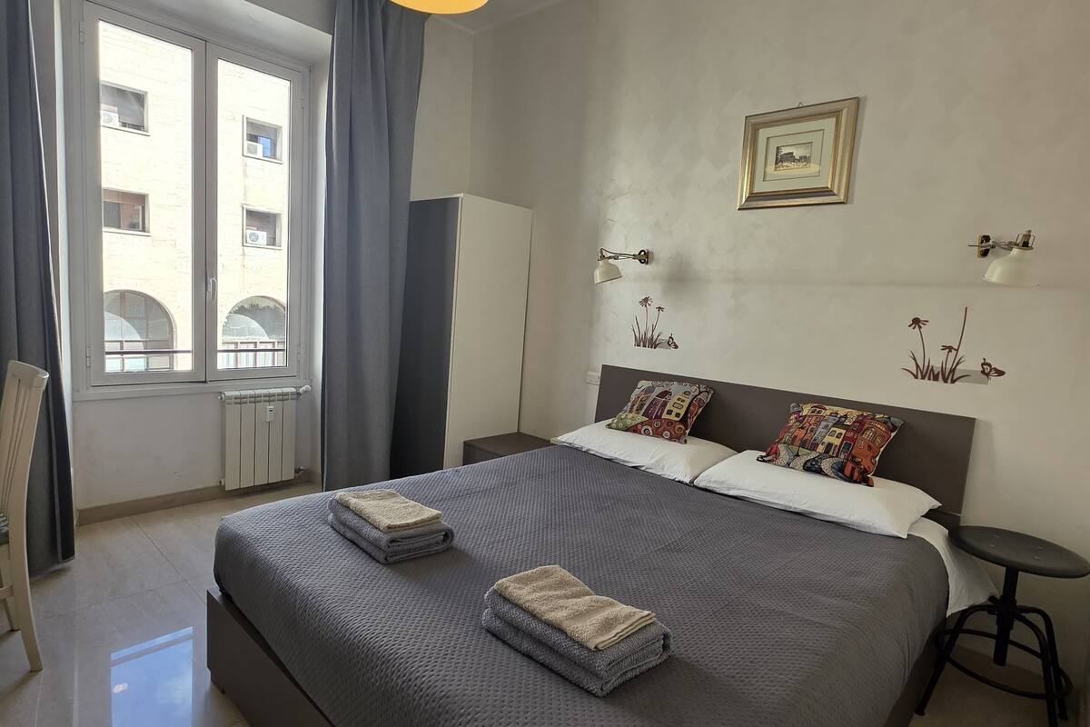 A neatly arranged hotel room with a large bed, a bedside table, a chair, a window with curtains, a radiator, and a ceiling light.