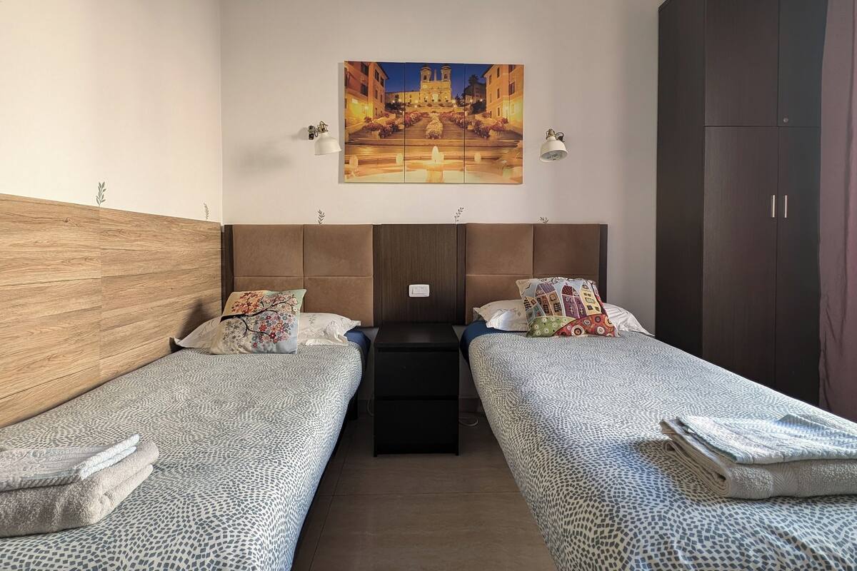 A room with two beds, a wooden headboard, a painting of a town square, and a wardrobe.
