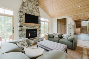 Ein geräumiges Wohnzimmer mit Kamin aus Stein, großen Fenstern, Holzboden und einem grünen Sofa.