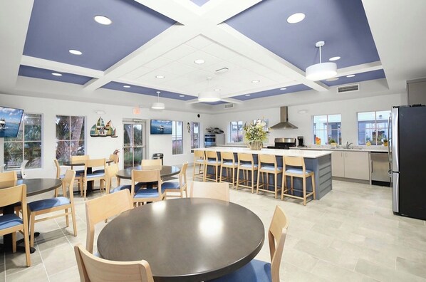 A modern dining area with round tables and chairs, a kitchen with a central island, and large windows providing views. - Coastal Retreat: Pool, Kayak, Beaches & Bikes (Jensen Beach)