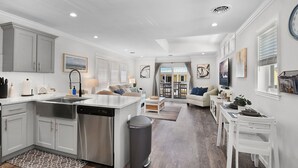 Una cocina moderna con electrodomésticos de acero inoxidable, un comedor con mobiliario blanco y una sala de estar con sofá y televisor.