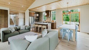 Een ruime woonkamer met een zithoek, een eettafel met stoelen, een keuken met een centraal kookeiland, en een groot raam met uitzicht op de omgeving.