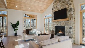 A spacious living room with a stone fireplace, large windows, and a wooden ceiling. - Refined Mountain Cabin on Pisgah Trails (Old Fort)