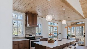 A modern kitchen with wooden beams, a central island with bar stools, and large windows. - Refined Mountain Cabin on Pisgah Trails (Old Fort)