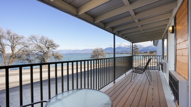 Una terraza de madera con una mesa y sillas de vidrio, con vista a un cuerpo de agua y montañas.