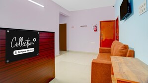 A reception area with a sign reading 'Collection Company-Serviced,' a wooden reception desk, a brown sofa, a wooden table, a flat-screen TV mounted on the wall, and a fire extinguisher. - Classic Havens Residency (Chengalpattu)