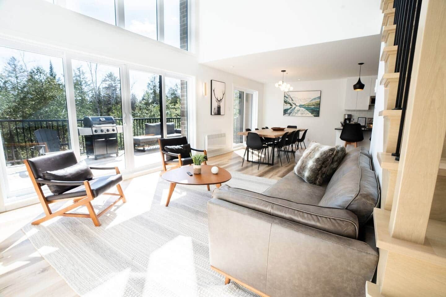 A spacious living room with a large sofa, a coffee table, and a dining area with chairs. There is a grill outside the large windows.