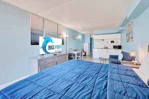 Ein modernes Hotelzimmer mit einem großen Bett, einem Schreibtisch, einem Sessel, einer Küchenzeile und einem Fernseher, der 'BIG FISH Rentals' anzeigt.