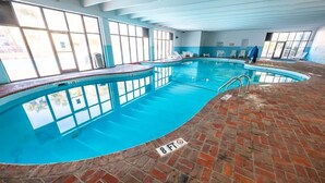 Una piscina interna con acqua cristallina e blu, circondata da pavimento in mattoni e ampie finestre.