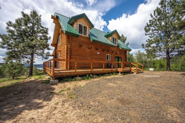 A log cabin with a wooden deck and a metal roof, surrounded by trees and a clear sky.