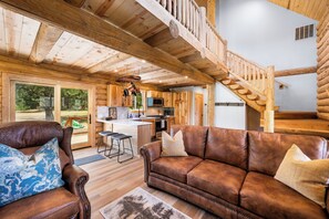 A log cabin interior with a wooden staircase, a living room with a brown leather sofa, and a kitchen area.