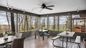 Une terrasse couverte avec une table et des chaises, un ventilateur de plafond, des guirlandes lumineuses et de grandes fenêtres offrant une vue sur les arbres et un bâtiment situé au loin.