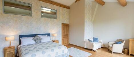 Un dormitorio con una cama grande, vigas de madera, un ventilador de techo, dos mesitas de noche, una silla y un mueble pequeño.