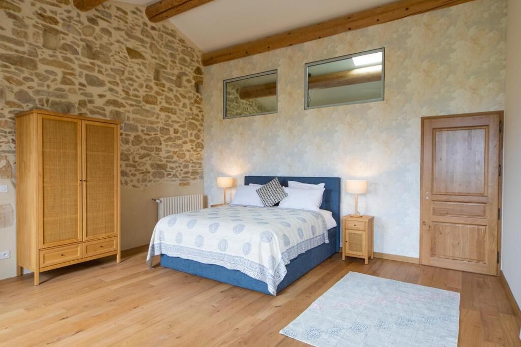 Une chambre avec un mur en pierre, une armoire en bois, un lit recouvert d’une courtepointe bleue, deux lampes de chevet et une porte en bois.