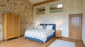 Une chambre avec un mur en pierre, une armoire en bois, un lit recouvert d’une courtepointe bleue, deux lampes de chevet et une porte en bois.