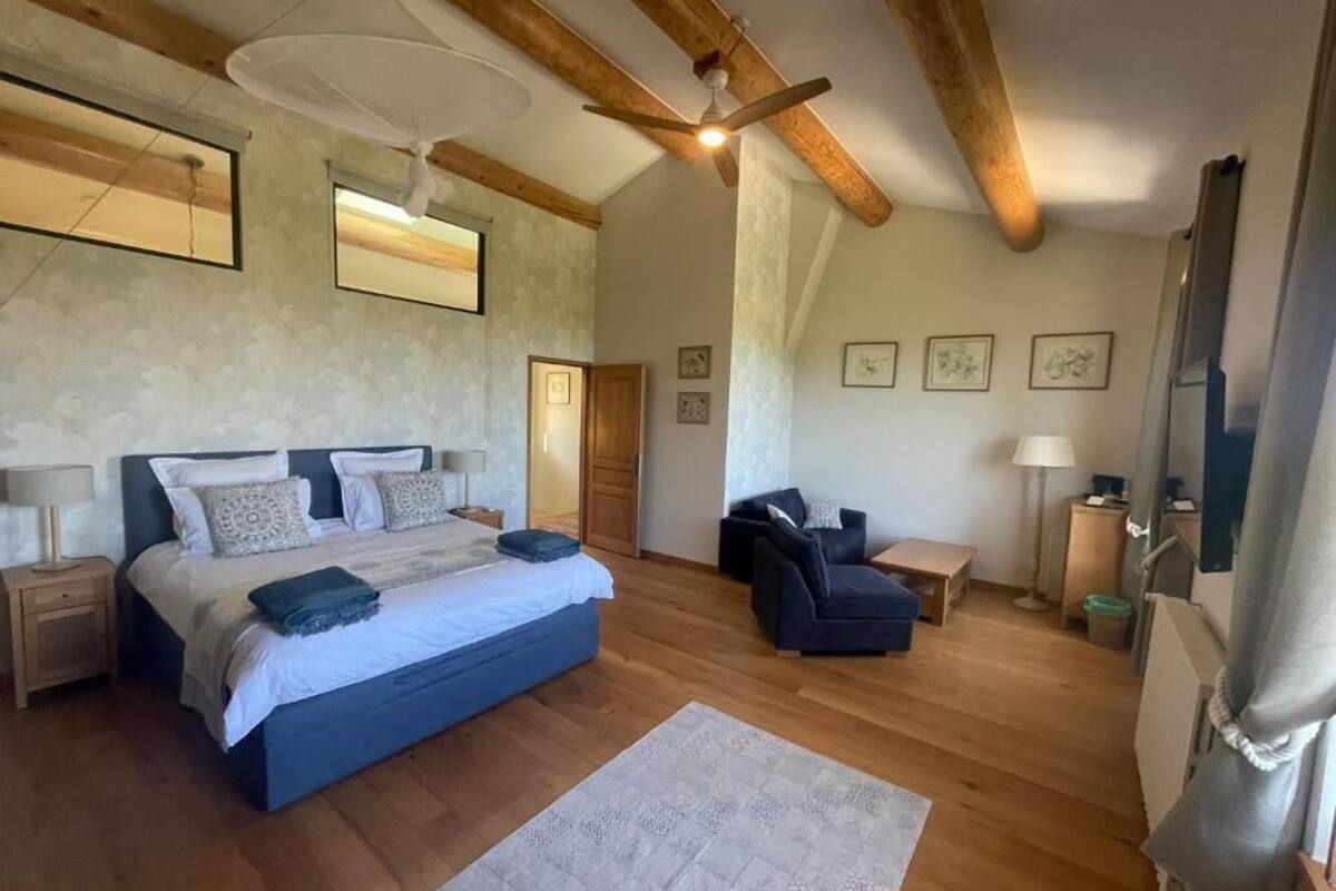 Un dormitorio con una cama grande, un sofá, un ventilador de techo, vigas de madera y cuadros en las paredes.