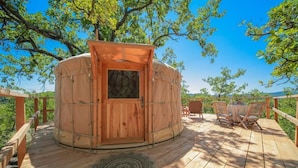 A wooden yurt with a door, a round table with chairs, and a patterned mat on a wooden deck surrounded by trees. - Les Yourtes de Provence (Saint-Paulet-de-Caisson)