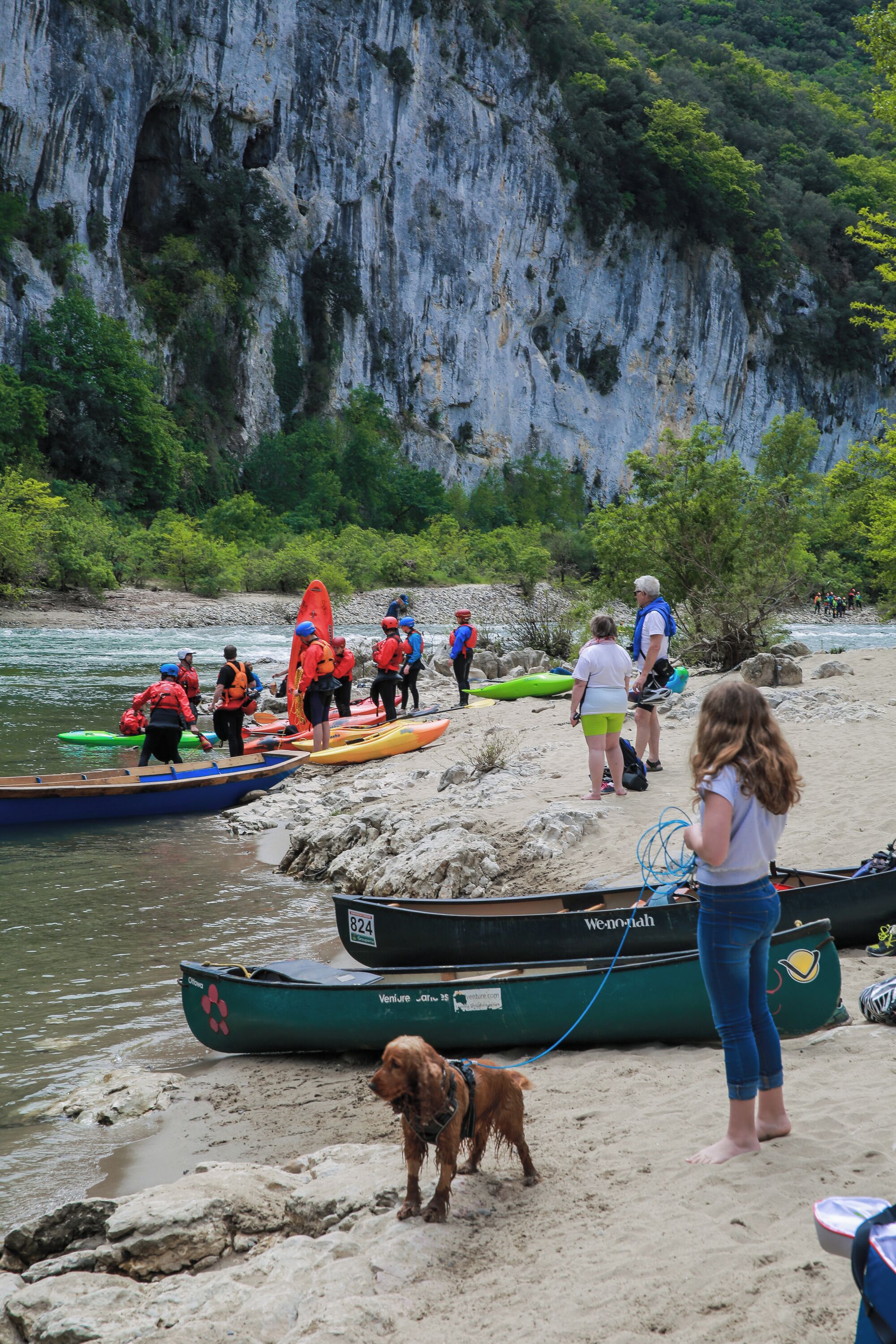 Un gruppo di persone prepara kayak su una spiaggia sabbiosa con una scogliera rocciosa sullo sfondo.
