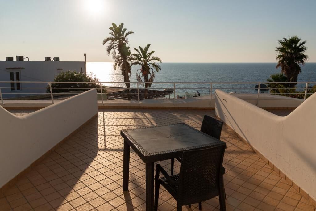 A terrace with a table and chairs, overlooking the sea with palm trees and a clear sky.