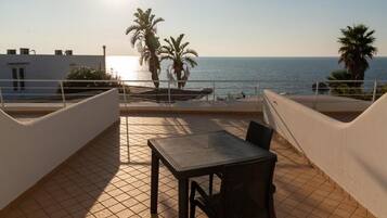 A terrace with a table and chairs, overlooking the sea with palm trees and a clear sky.