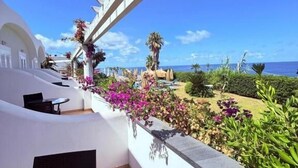 A balcony with a view of the sea, surrounded by white walls and tropical plants.