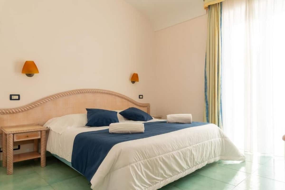 Una cama bien hecha con ropa de cama azul y blanca, dos lámparas fijadas a la pared y una ventana con cortinas.