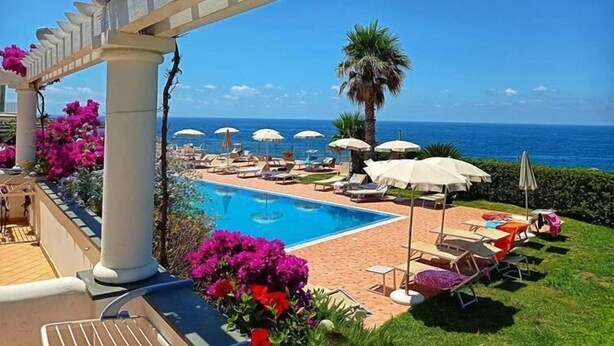 A poolside area with lounge chairs and umbrellas, a clear view of the ocean, and a pergola with flowers.
