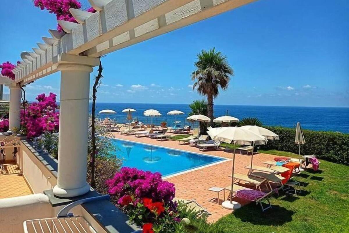 Zona junto a la piscina con sillas de descanso y sombrillas, vista despejada del océano, y una pérgola adornada con flores.