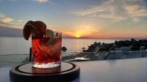 A glass of red drink with a lemon slice on a coaster, overlooking a sunset over the sea with buildings in the background.