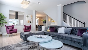 Una sala de estar moderna con un sofá gris, dos sillones morados, una mesa de centro redonda y una escalera.