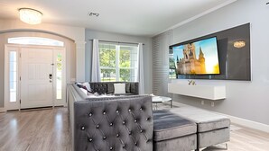 Una sala de estar moderna con un televisor de pantalla plana grande montado en la pared, un sofá gris con capitoné y un amplio ventanal con cortinas.