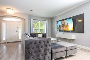 Ein modernes Wohnzimmer mit einem großen Flachbildfernseher an der Wand, einem grauen, gepolsterten Sofa und einem großen Fenster mit Vorhängen.