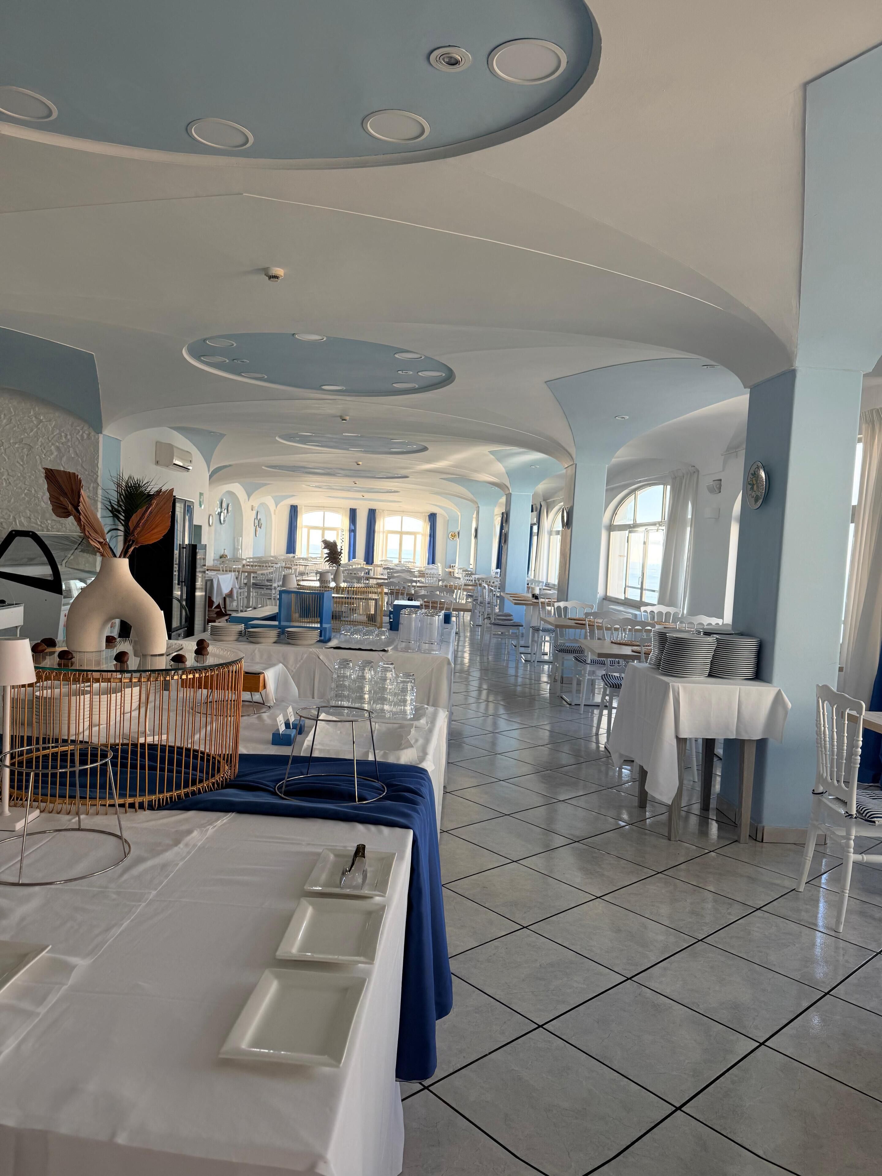 A spacious dining area with multiple tables set for service, a modern ceiling design, and large windows allowing natural light.