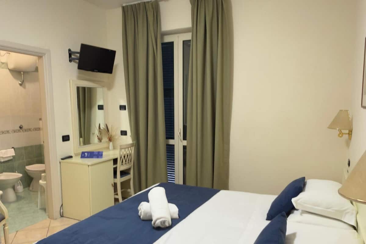 Une chambre d’hôtel comprenant un lit, un bureau avec un téléviseur à écran plat, une fenêtre avec des rideaux, une salle de bain avec des toilettes et un lavabo, et un téléphone sur la table de chevet.