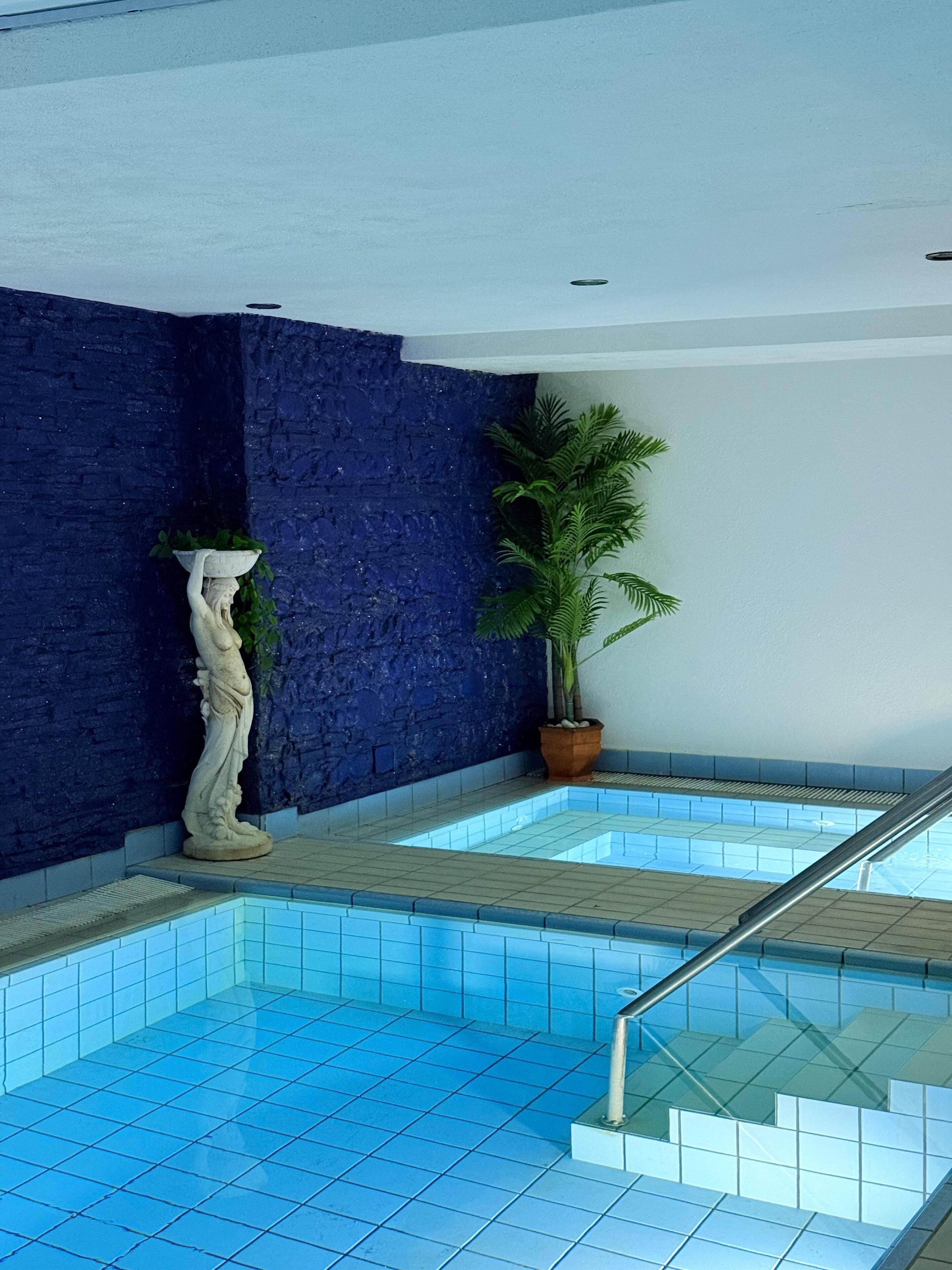 Une piscine aux carreaux bleus, une statue blanche, une plante en pot et un mur bleu à texture.